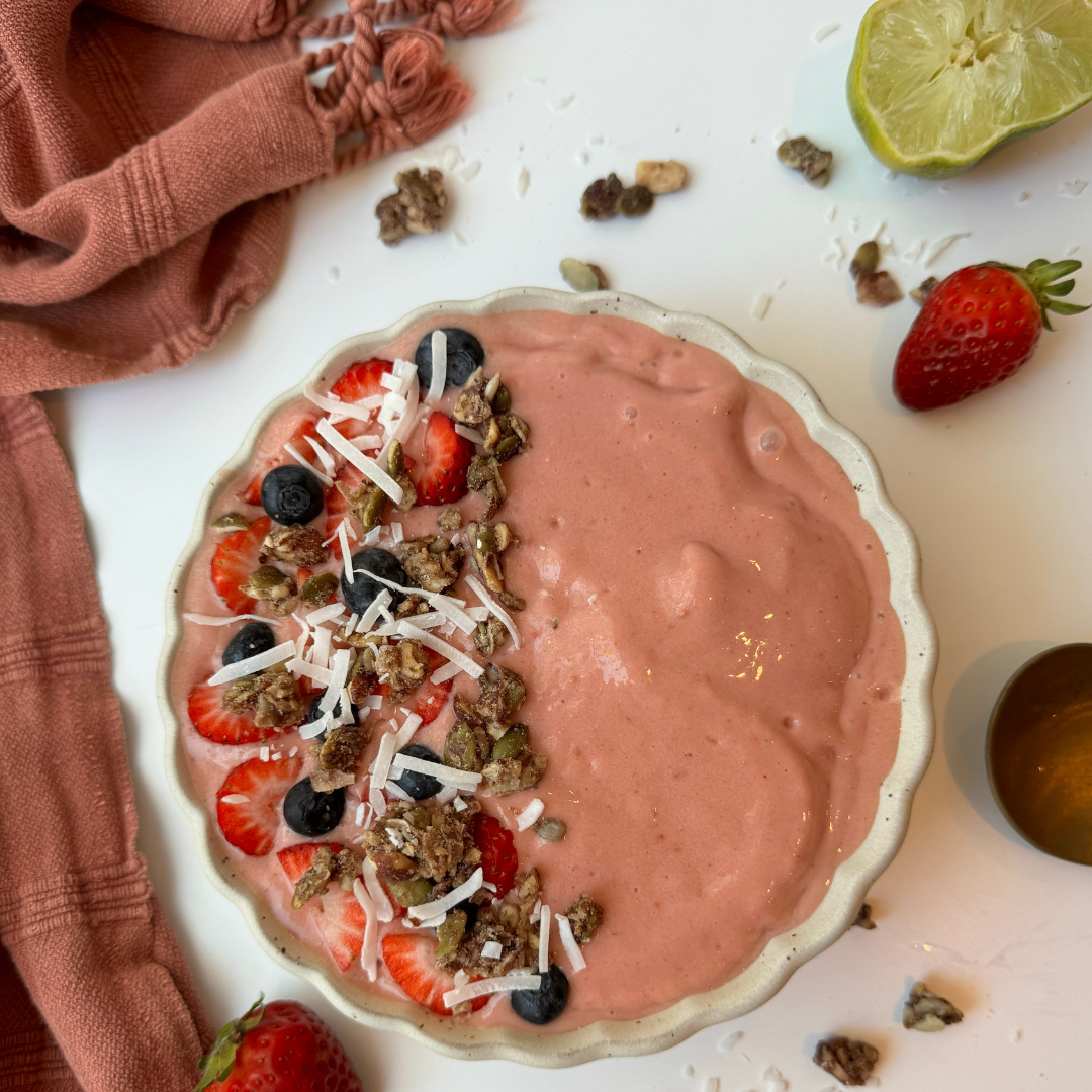 Pink tropical smoothie bowl topped with sliced strawberries blueberries coconut flakes and granola on a white table beside a pink scarf.
