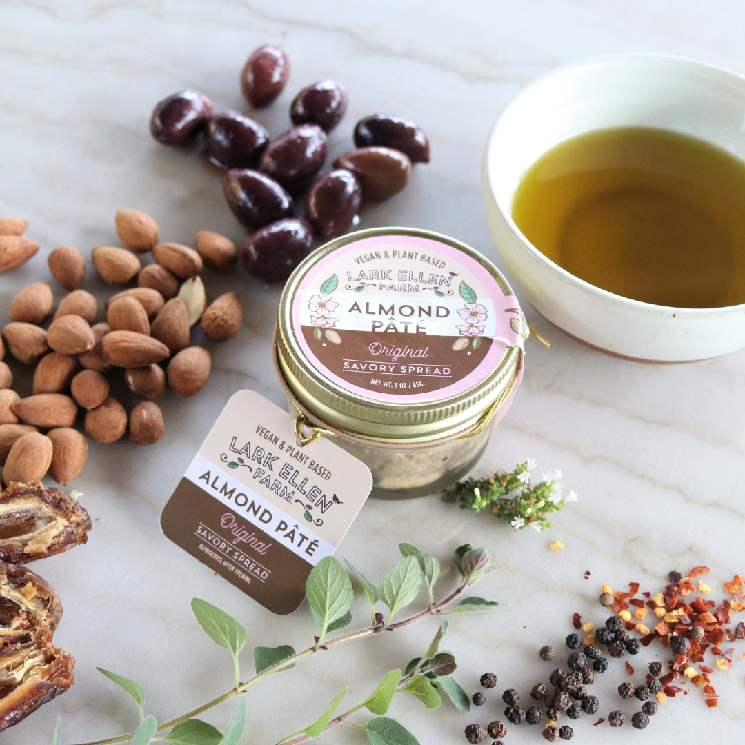Lark Ellen Farm almond pate jar surrounded by almonds olives and fresh herbs on a marble surface.