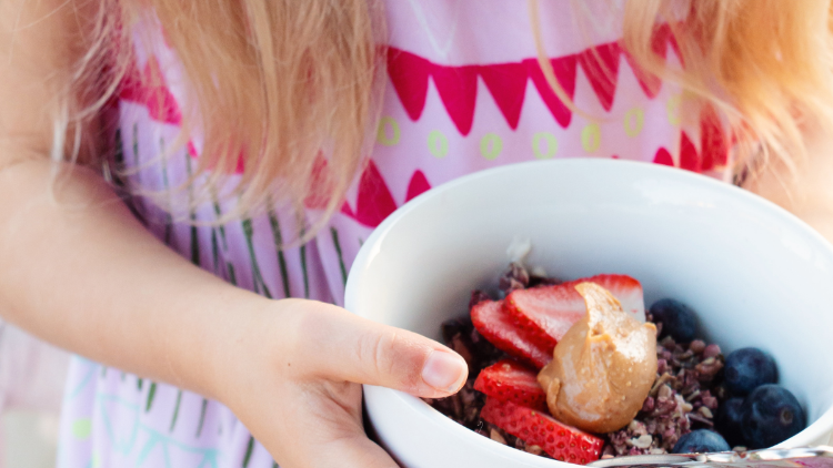 Child holding a white bowl of fruit and granola with strawberries blueberries and a dollop of peanut butter
