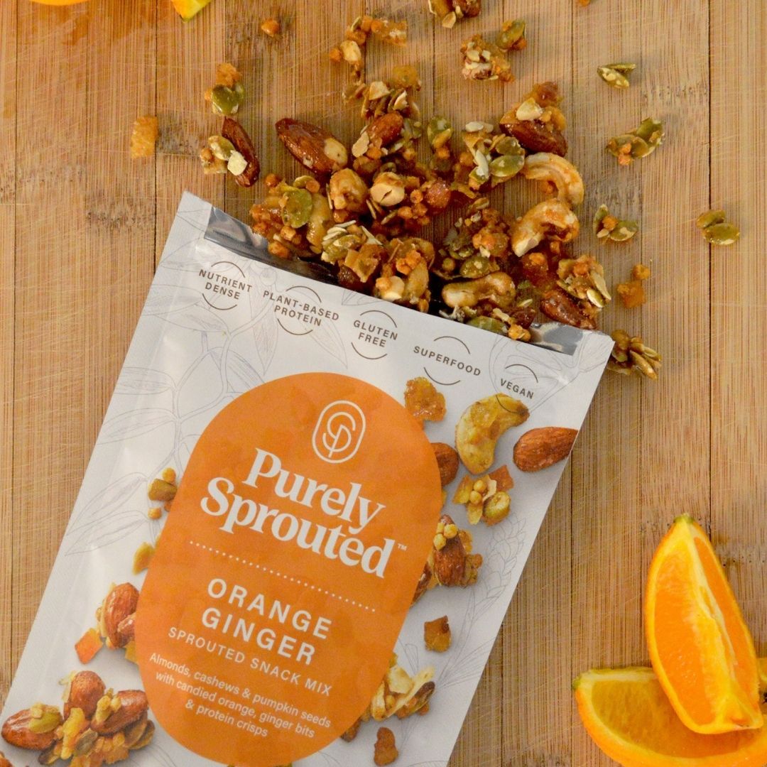 Open Purely Sprouted Orange Ginger snack mix bag with mixed nuts and seeds spilled on a wooden surface.