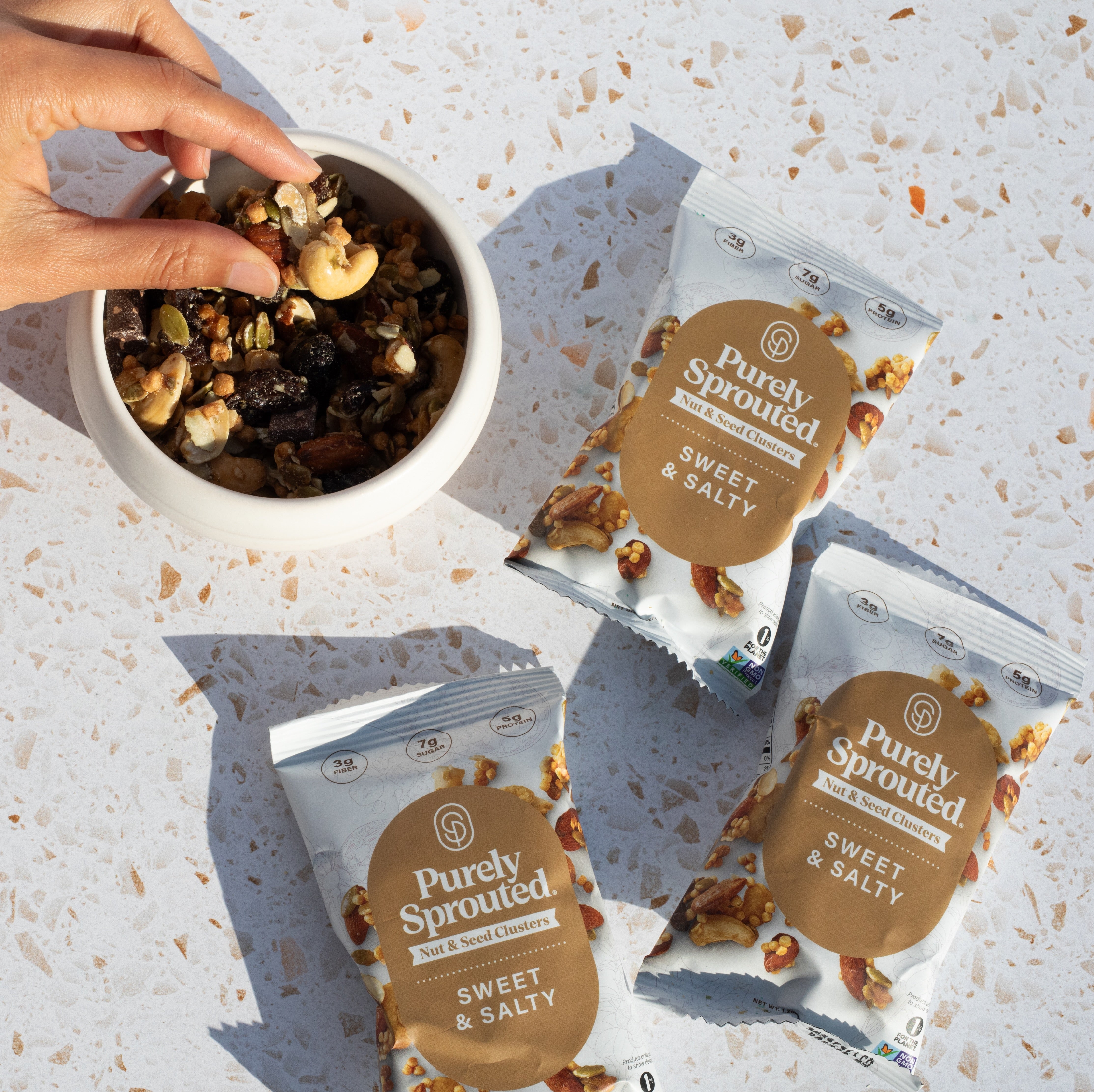 Hand scooping sprouted snack mix from a white bowl beside Purely Sprouted Sweet and Salty snack packs.