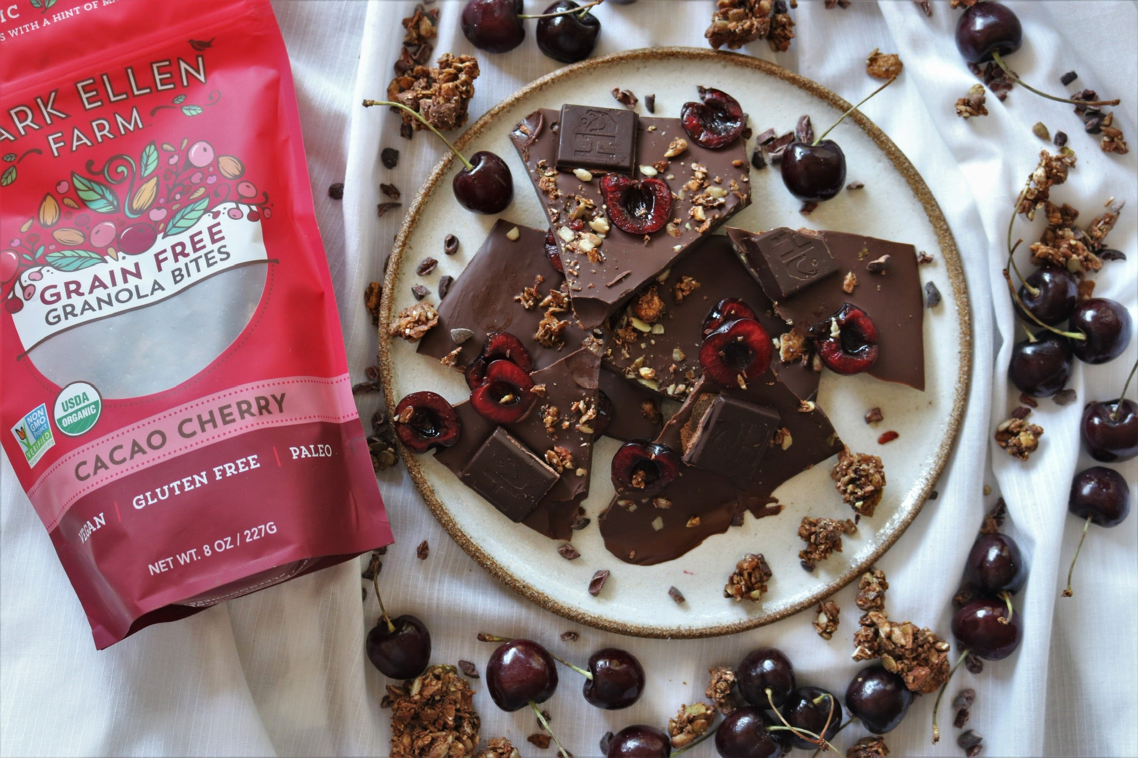 Lark Ellen Farm cacao cherry granola bag beside a plate of chocolate bark with cherries and granola clusters