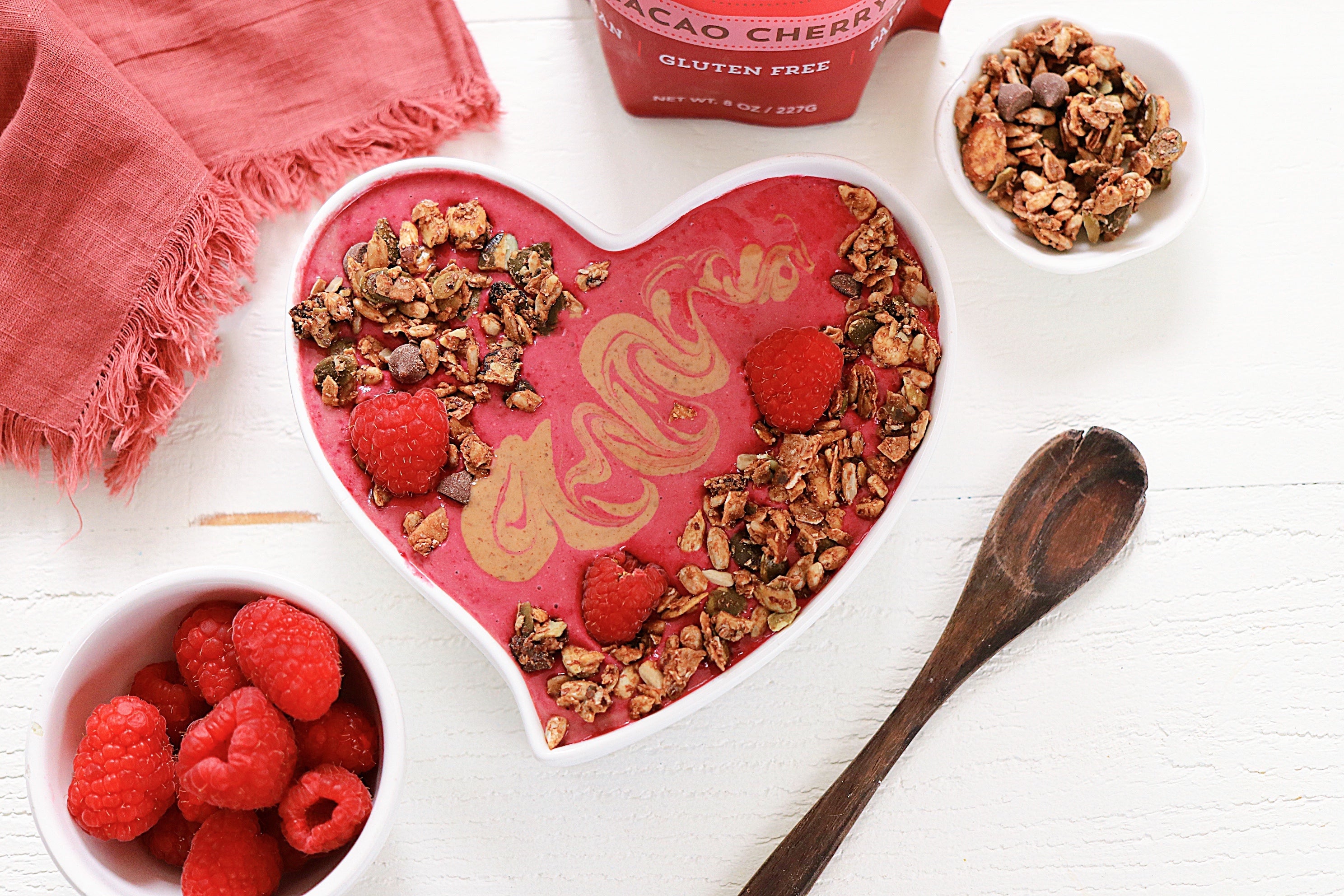 Heart shaped bowl of pink smoothie topped with granola and raspberries with the cacao cherry granola bag nearby