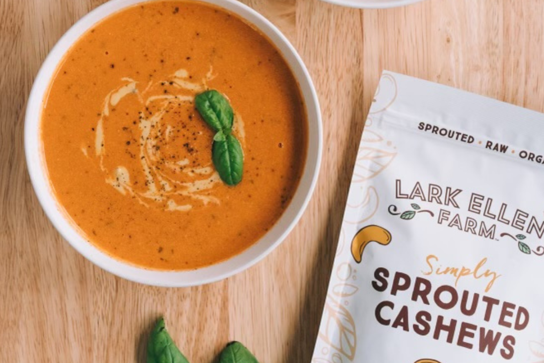 Creamy orange cashew soup in a white bowl with basil garnish beside a sprouted cashews bag on a wooden surface