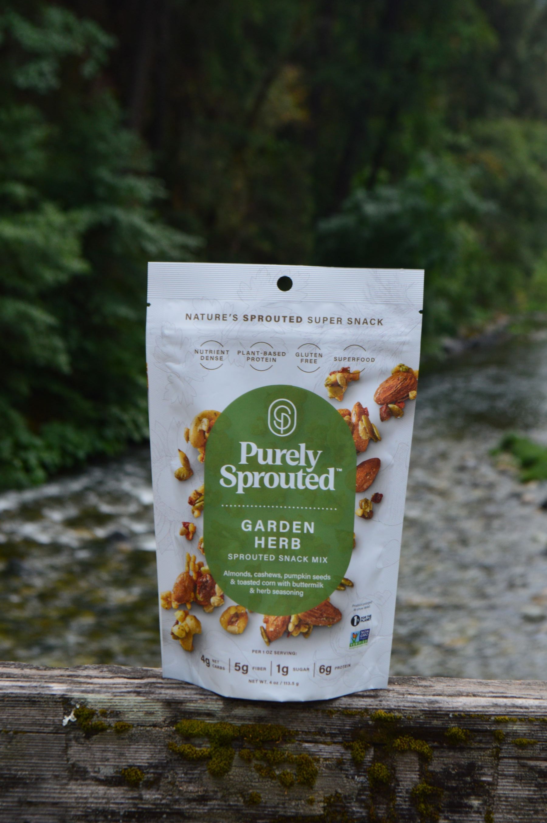 Garden Herb Sprouted Snack Mix bag displayed outdoors on a wooden railing beside a forest stream.