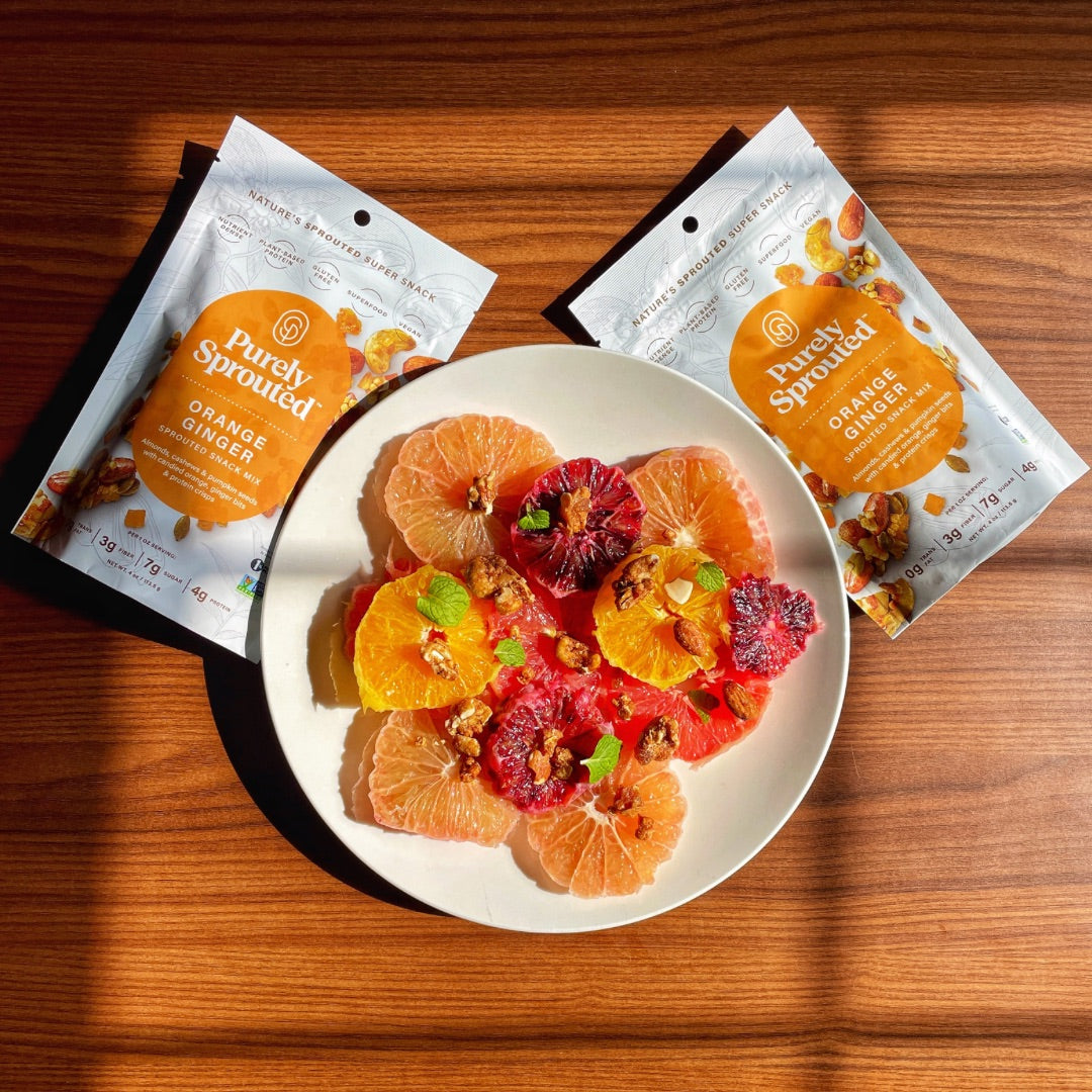 Two Purely Sprouted Orange Ginger snack mix pouches on a wooden table beside a plate of citrus slices and nuts.