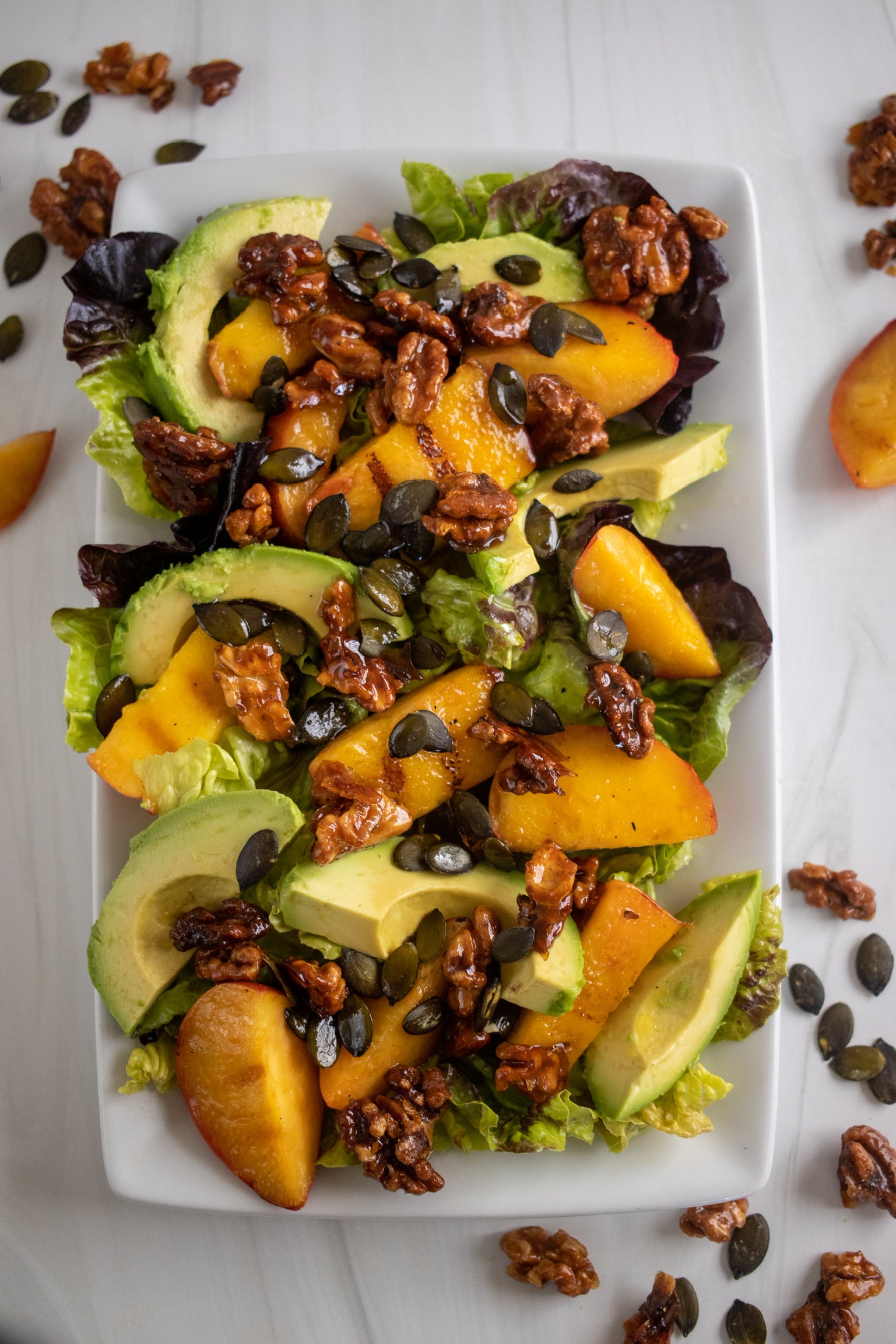 Fresh salad with mixed greens, avocado, peaches, walnuts, and pumpkin seeds on a white plate.