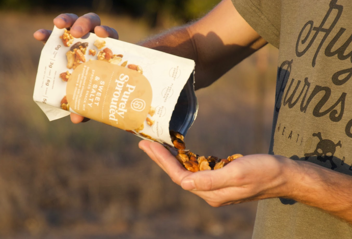 Person outdoors pours Sweet & Salty Sprouted Snack Mix from a white bag.